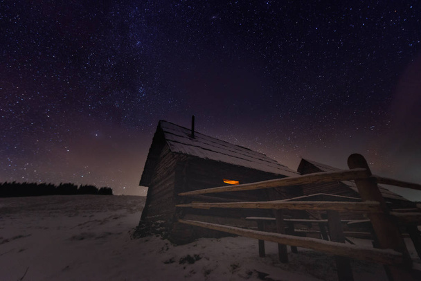 Величественный Млечный Путь над зимним горным ландшафтом. Ночная сцена. Деревянный дом со светом в окне. Кукульский хребет, Карпаты, Украина, Европа. - Фото, изображение