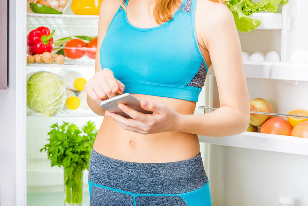 Молодая женщина, используя приложение для смартфона и оставаясь рядом с холодильником, полным здоровой пищи
.  - Фото, изображение