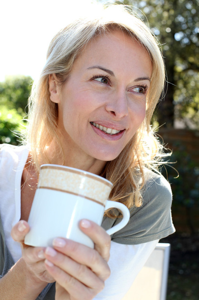 Portrait of blond woman with tea mug sitting outside - 写真・画像