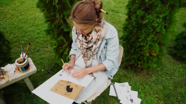 Nainen Illustrator hahmotellaan paikka Sketch paperille ja käyttämällä kyniä ja pitää kädessä. Tyttö istuu puistossa ulkona päiväsaikaan
. - Materiaali, video