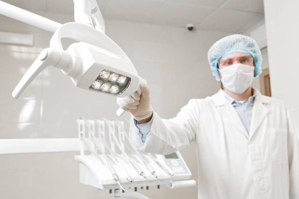 Visão inferior de um médico dentista retrato, cirurgião de uniforme e máscara em seu rosto, usando óculos, segurando lâmpada em sua mão
. - Foto, Imagem