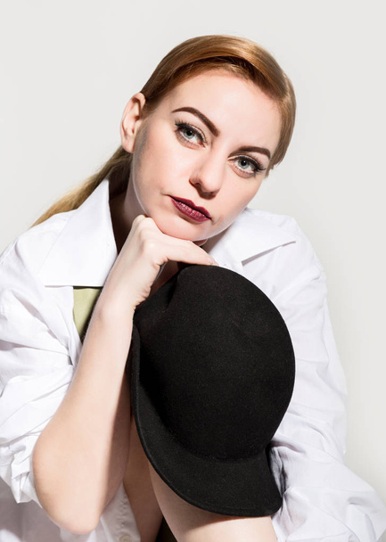naked young woman in a mans white shirt with green tie holding black hat and posing on the floor - Foto, afbeelding
