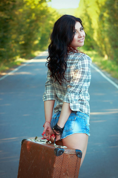 lonely brunette girl smiling and holding an old suitcase in the middle of the road - Fotografie, Obrázek