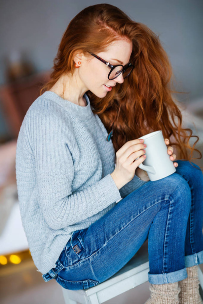 young red haired girl holding cup of tea sitting in Christmas decorated room - Foto, afbeelding