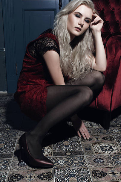 Beautiful blonde woman with hair and make-up sitting on the floor near the red chair - Foto, immagini