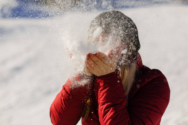 Picture of attractive teenage girl blowing snow on her palms while wearing winter jacket with bokeh background - Photo, Image