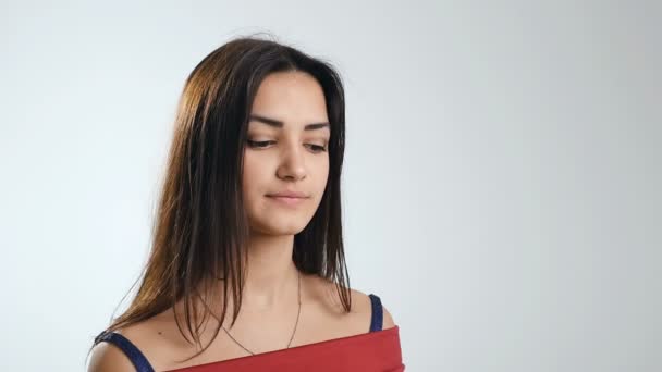 Een mooie 18-jarige meisje in een rode jurk met open schouders glimlacht raadselachtig en deelt haar positieve emoties terwijl ze in de studio, in slo-mo - Video