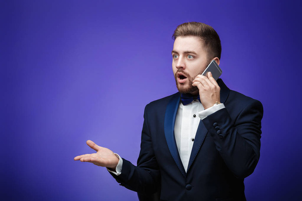 successful man in tuxedo and bow tie talking on phone. businessman holding smartphone, blue background - Foto, afbeelding