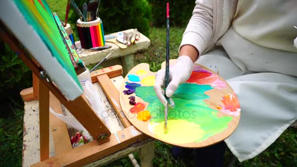 Europees uiterlijk kunstenaar houdt palet en penselen, vrouwelijke Meng gele kleur met een andere kleur en maakt eerste slagen op Canvas in openlucht parkeren. - Video