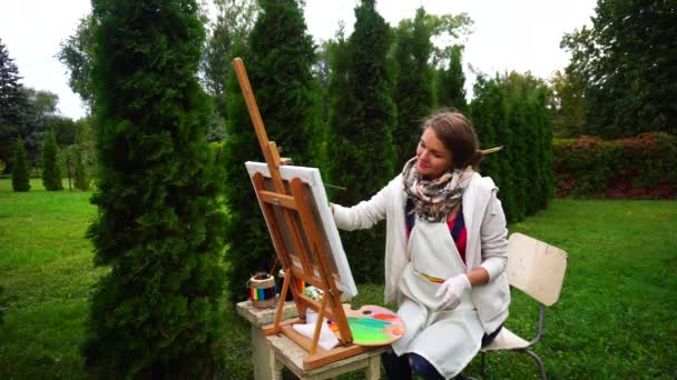 Impresionante artista mujer sentada en el caballete, dibuja en el parque en el fondo de árboles de coníferas al aire libre
. - Imágenes, Vídeo