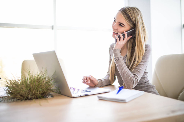 Portret van een jonge zakenvrouw spreken op de telefoon en met behulp van laptop op kantoor - Foto, afbeelding