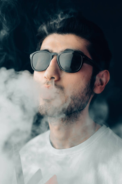 Young cool guy in sunglasses exhales a cloud of smoke. Vertical studio portrait in close-up. - Fotografie, Obrázek