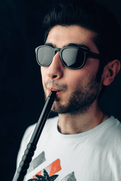 Young guy with glasses inhales through the mouthpiece of a hookah. Vertical close-up portrait. - Fotoğraf, Görsel