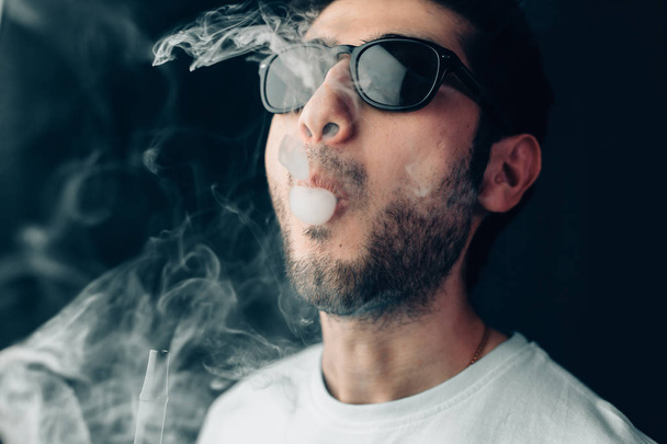 Nuori, siisti mies aurinkolaseissa hengittää ulos savupilven. Studio vaakasuora muotokuva lähikuva
. - Valokuva, kuva
