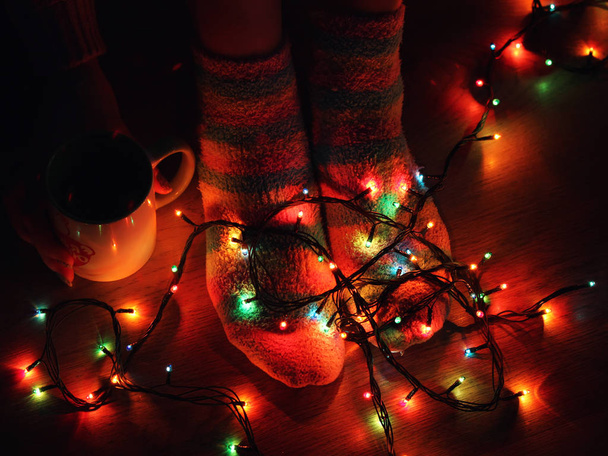 Female feet close up at home in warm socks on which is multi-colored garland and near hand with a cup of hot tea - Photo, Image