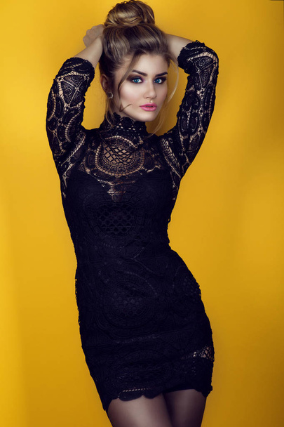Portrait of a gorgeous young glam lady with updo hairstyle wearing tight-fitting short black lace dress and stockings. Isolated on bright yellow background.  - 写真・画像