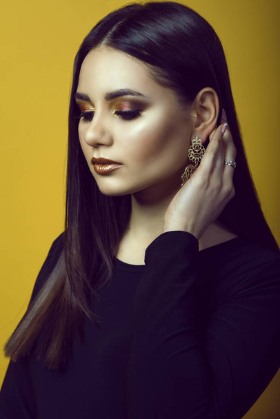 Portret van jonge mooie dark-haired meisje met professionele make-up in gouden en koperen kleuren verbergen van haar haar achter het oor met luxe lange oorbel. - Foto, afbeelding
