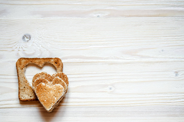 На белой деревянной поверхности лежит тост с иссечением в сердце. Сверху на тосте лежат сердца хлеба
. - Фото, изображение