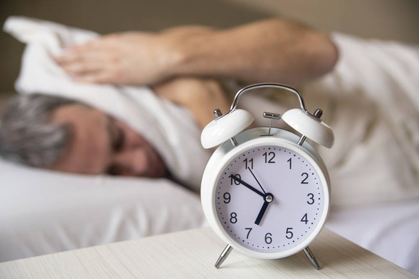 sleeping man disturbed by alarm clock early morning.  Sleepy young man covering ears with pillow as he looks at alarm clock in be - Photo, Image