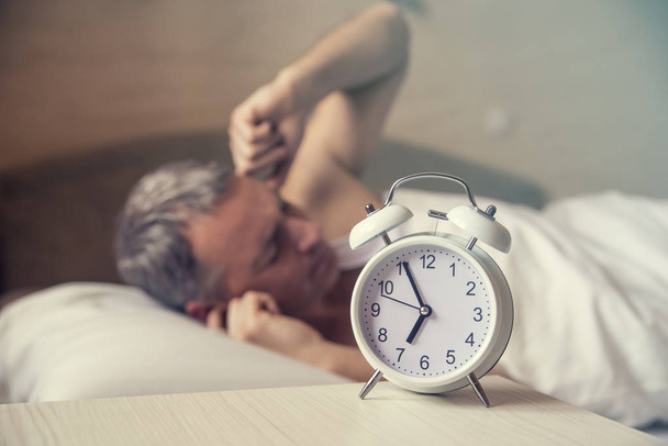 sleeping man disturbed by alarm clock early morning. Angry man in bed awoken by a noise. Waked Up. Man lying in bed turning off an alarm clock in the morning at 7am - Фото, зображення