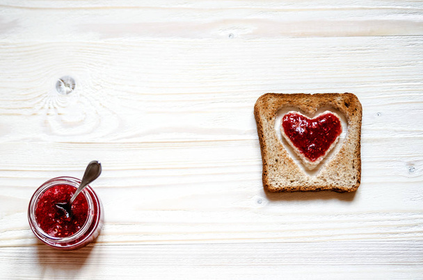 Toasted white bread with a heart inside. At the heart plastered with raspberry jam. To the left of French toast on a white surface is a jar of jam, a jar of sticks dessert spoon. - Photo, Image