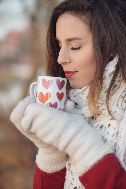 Ritratto ravvicinato di una bella giovane donna caucasica sorridente in giacca e sciarpa invernale, che tiene una tazza di caffè o tè, all'aperto nel parco o nella foresta durante la giornata invernale. Modo invernale
 - Foto, immagini