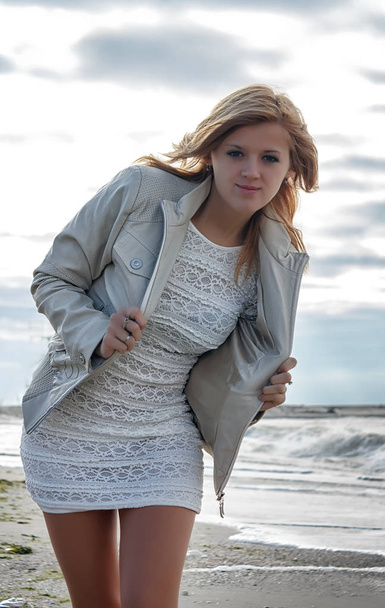 Молодая блондинка в белом платье в драпированной куртке позирует на песчаном пляже у моря
 - Фото, изображение