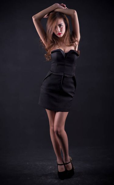 Hermosa mujer sexy con cuerpo delgado perfecto y pelo rizado largo en vestido negro posando sobre fondo negro. Captura de estudio
. - Foto, Imagen