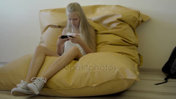 Teenager girl sitting on armchair and watching video on smartphone - Materiał filmowy, wideo