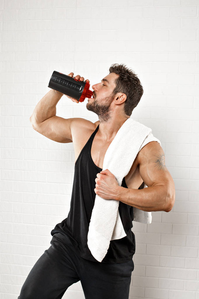 Handsome sportsman standing with towel holding a bottle of gainer while drinking over white background. - Photo, image