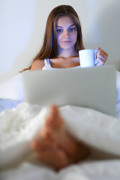 Pretty young woman with laptop in bed and enjoying a cup of tea - Fotó, kép