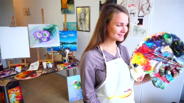 Mulheres belo pintor posando e sorrindo na câmera com pincéis e paleta no estúdio de arte pendurado com esboços e desenhos
. - Filmagem, Vídeo