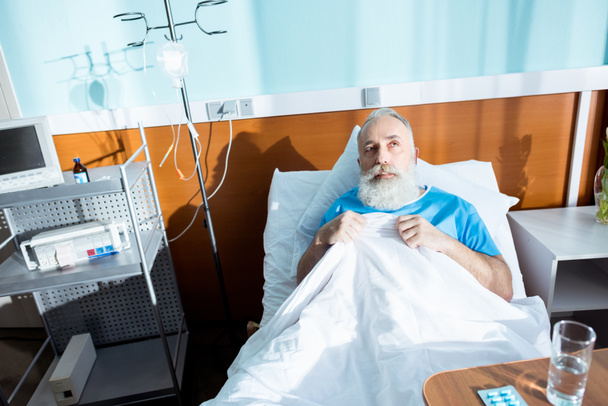 Хворий старший чоловік лежить в лікарняному ліжку і коригує ковдру
 - Фото, зображення