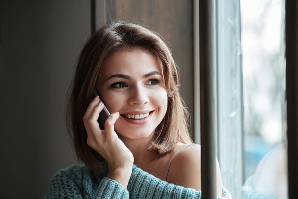 Cheerful young lady sitting near window talking by phone. - Фото, зображення