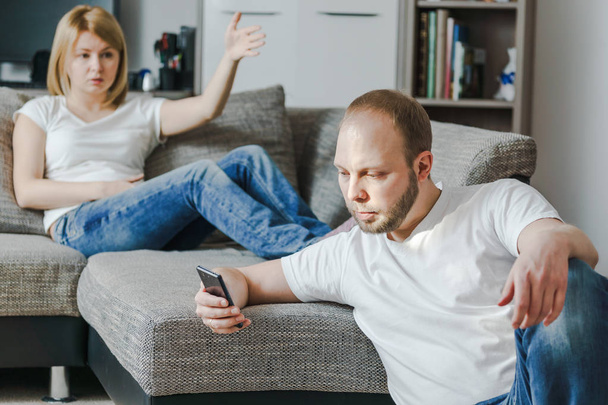 Mujer joven enojada sentada en el sofá hablando con su marido mientras él está usando su teléfono celular en su sala de estar
. - Foto, imagen