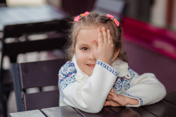 Эмоциональная девушка за столом в кафе. Милая маленькая девочка сидит в ресторане на открытом воздухе в летний день. ребенок в кафе ждет своего заказа
.  - Фото, изображение