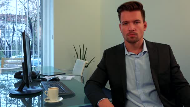 A young, handsome man sits at a table in an office and looks at the camera - Video