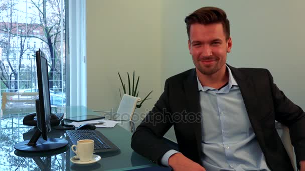 ein junger, gutaussehender Mann sitzt an einem Schreibtisch in einem Büro, nickt und lächelt in die Kamera - Filmmaterial, Video