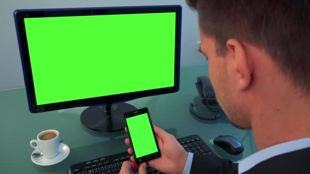 Un uomo si siede in un ufficio e guarda a turno uno schermo del computer e uno smartphone (entrambi schermo verde
) - Filmati, video