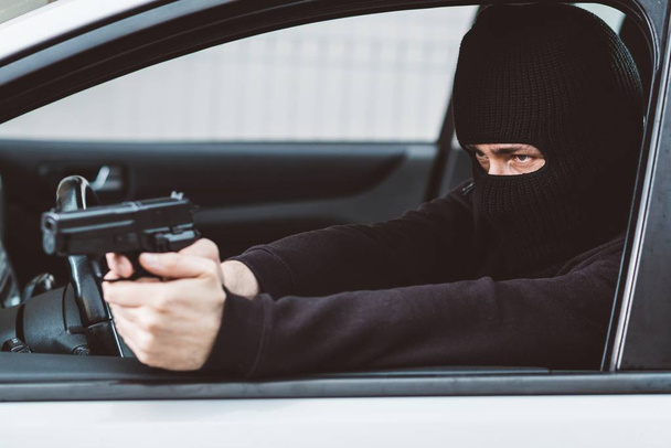 Homme en cagoule noire avec arme de poing conduisant une voiture
. - Photo, image