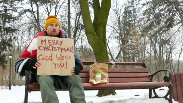  4 k. άστεγος στο χειμερινό πάρκο της πόλης με χαρτόνι καλά Χριστούγεννα - Πλάνα, βίντεο