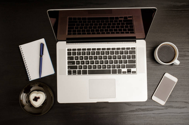 Vista superior de un teclado portátil, taza de café, un teléfono inteligente, un cuaderno y un postre en una mesa de madera negra
 - Foto, Imagen