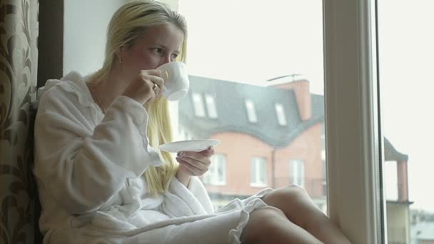 Mädchen im Bademantel trinkt Kaffee zu Hause am Fenster - Filmmaterial, Video