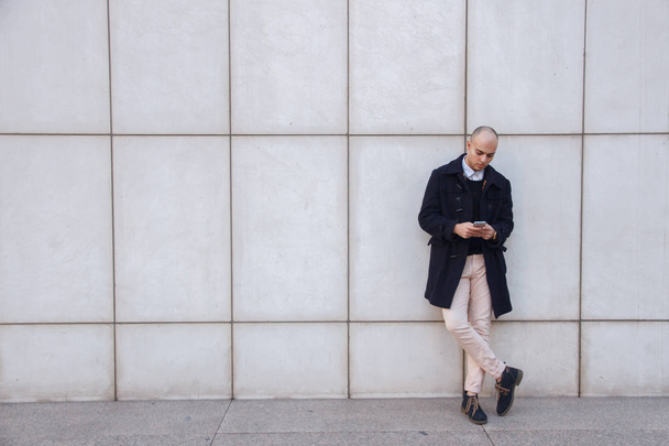 Joven hombre de negocios calvo guapo sosteniendo un teléfono inteligente, mirando hacia abajo golpeando la pantalla apoyada en una pared blanca
.  - Foto, Imagen