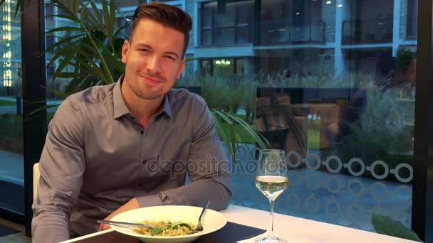 Egy fiatal, jóképű férfi ül egy asztalnál, egy étterem és mosolyog a kamera, ételt és pohár bort az asztalon - Felvétel, videó