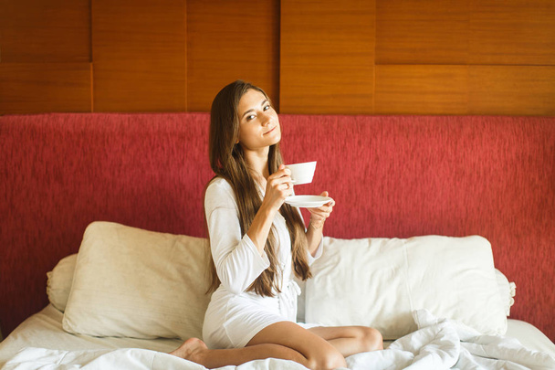 young girl in white robe drinking tea in bed - Фото, зображення