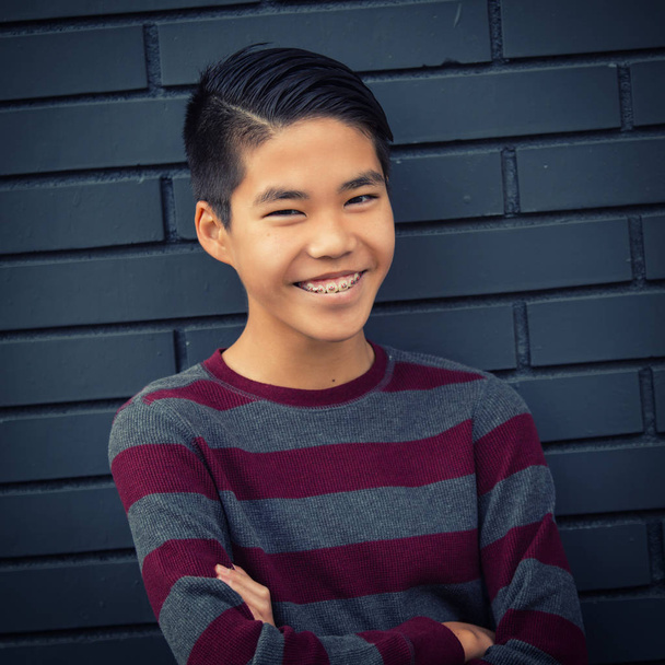Πολύ χαρούμενος εφηβικό ασιατικό αγόρι με τιράντες, χαμογελώντας με τα χέρια διπλωμένα - Φωτογραφία, εικόνα