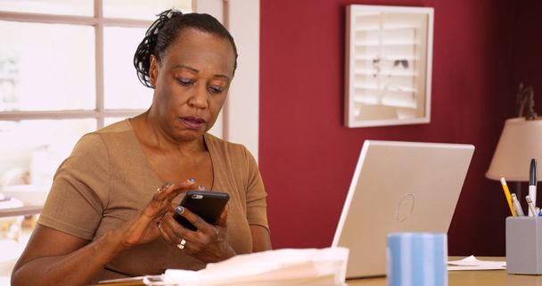 Vanhempi musta nainen käyttää puhelintaan ja kannettavaansa veroissaan.
 - Valokuva, kuva
