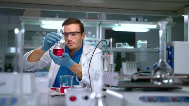 Chemik studia płynem w szklanej kolbie w nowoczesnym laboratorium. Pracownik laboratorium - Materiał filmowy, wideo