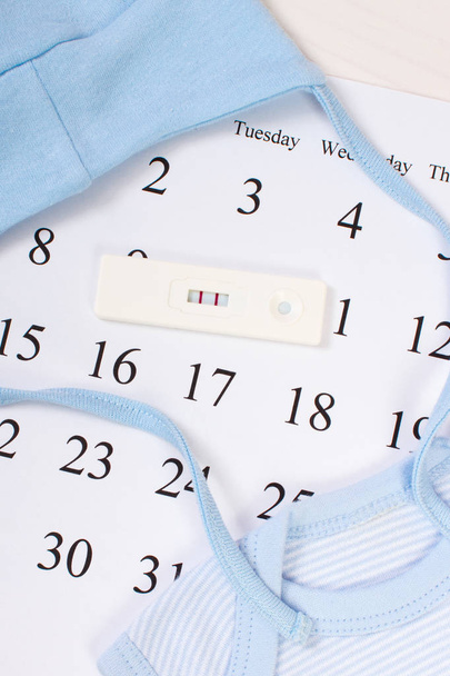Zwangerschapstest met positief resultaat en kleding voor pasgeboren op kalender, verwacht voor baby - Foto, afbeelding